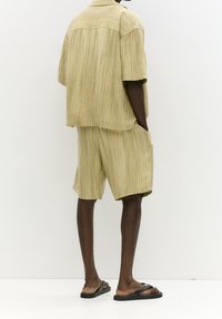 Licht khaki korter-mouw overhemd en bijpassende shorts in een gestructureerde stof, met subtiele verticale strepen, gecombineerd met zwarte slippers.