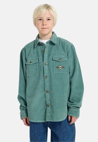 Groene corduroy button-up jas met twee borstas, bruine knopen en een ruitvormige patch op het linker borstgebied.