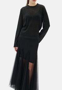Femme portant un haut noir transparent pailleté à manches longues avec une jupe noire comportant des panneaux en maille transparente superposés.