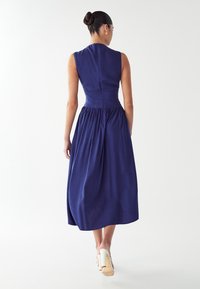 Haljina bez rukava, do koljena, u tamnoplavoj boji s uskim korzetom i nabranom suknjom. Zatvaranje na patentni zatvarač straga i glatka tekstura.