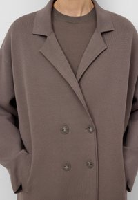 Cappotto marrone doppiopetto con ampi revers indossato sopra un maglione marrone a girocollo abbinato, mani nelle tasche del cappotto, vista ravvicinata.