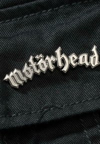 Metallpin som har ordet "Motörhead" i fet, sølvfarget gotisk skrift, festet til en teksturert svart stoffbakgrunn.