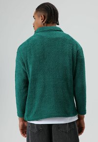 Groene fleecejas met een gestructureerde afwerking, lange mouwen en een omgeslagen kraag. Heeft een iets langere achterkant voor extra comfort.