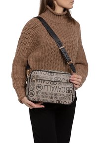 Borsa a spalla beige con logo Cavalli neri. Forma rettangolare, doppi scomparti con cerniera e tracolla nera regolabile con dettagli del logo.