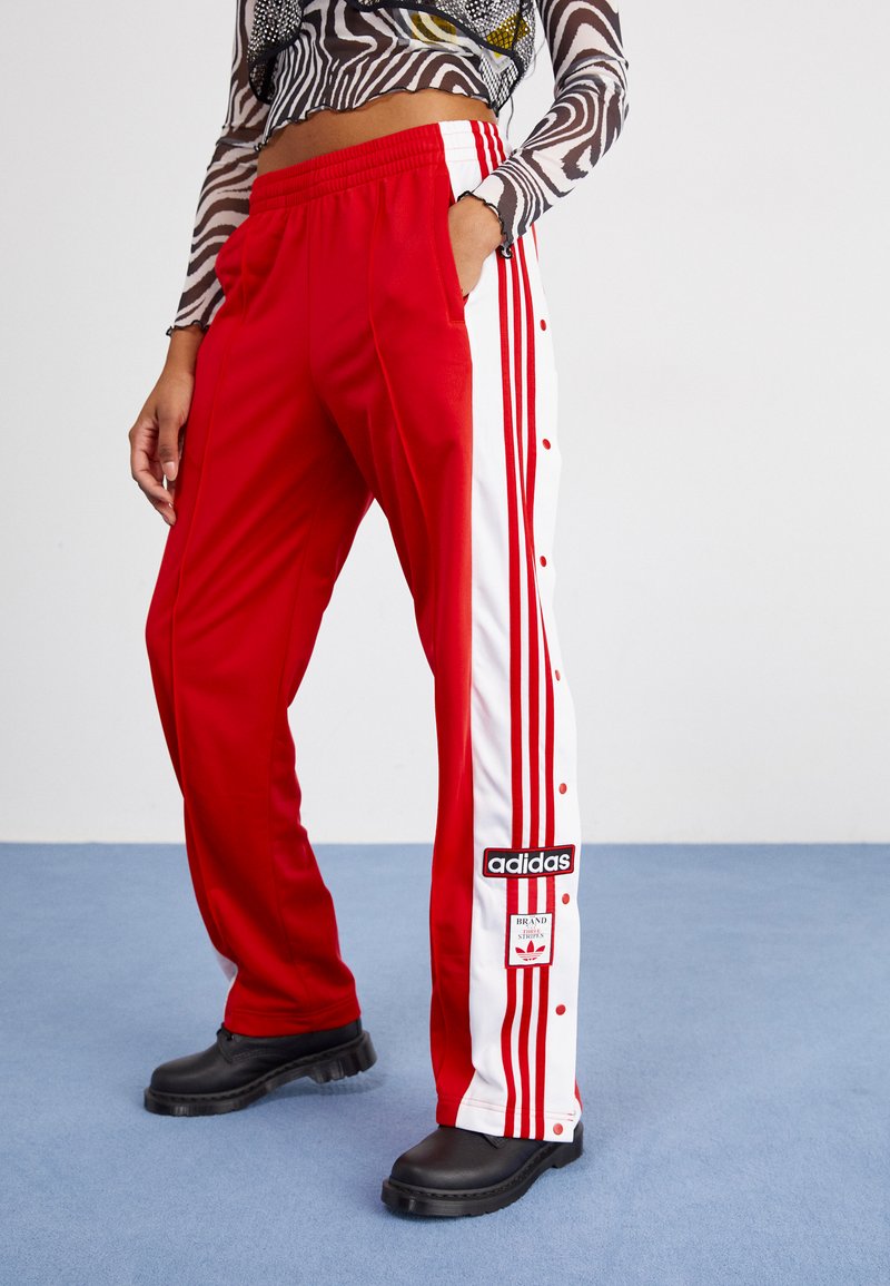 Calças atléticas vermelhas com riscas brancas nas laterais, botões de pressão e um patch do logo Adidas. Feitas de um material suave e flexível, com uma cintura elástica.