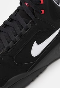 Černá atletická obuv s hladkým syntetickým svrškem, bílým logem, texturovanými akcenty, černými tkaničkami a červenými nýty. Vyztužená špička a gumová podrážka.