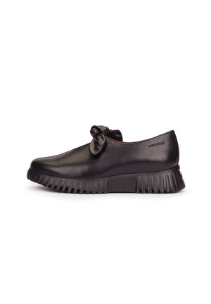 Zapato de mujer de cuero negro con suela gruesa y estriada y un nudo decorativo de cuero en la parte superior, vista lateral sobre fondo blanco.