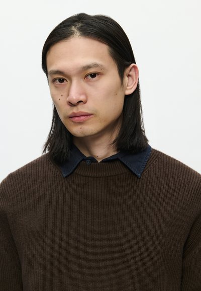 Pulôver castanho de malha canelada com um decote estruturado, usado sobre uma camisa de colarinho azul-escuro. A pessoa tem cabelo preto liso e comprido e uma expressão neutra.