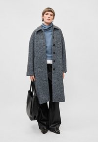 Vilnas maisījuma mētelis ar herringbone rakstu, pelēkās un zilās krāsās, ar lieliem pogām un apkakles izgriezumu. Melna soma, plati svārki un uzsistie apavi.