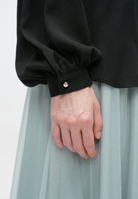 Čierna blúzka s nafúknutými rukávmi a manžetou so zlatými gombíkmi. Ruka drží svetlomodrý tylový sukňu, ktorý ukazuje jemnú textúru.