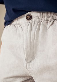 Jasnoszare lniane spodnie z elastycznym pasem i brązowym guzikiem jako akcentem. Tkanina ma subtelną fakturę.