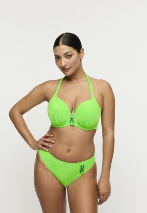 Vrouw met donker haar in een felgroene halterbikini met decoratieve ringen, staat zelfverzekerd met één hand op haar heup tegen een witte achtergrond.