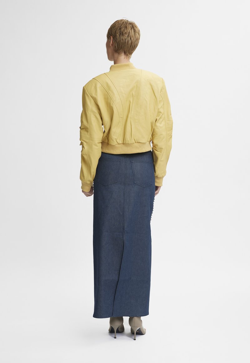 Veste bomber en cuir jaune avec des coutures texturées, associée à une longue jupe en denim, avec une coupe droite et un détail fendu à l'arrière.