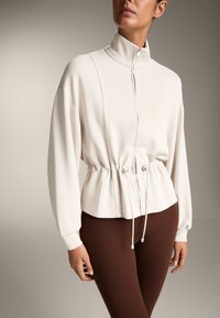 Lys beige zippered jakke med høj krave, samlet talje og lange ærmer, parret med mørkebrune leggings. Glat stofstruktur.