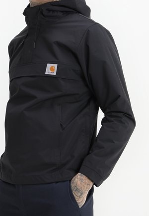 Osoba ubrana w czarną kurtkę z kapturem z przednią kieszenią i pomarańczowym logo Carhartt, ręka w kieszeni, pokazująca tatuaż na nadgarstku.