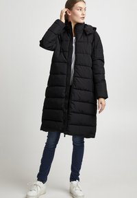 Doudoune noire avec capuche, fermeture éclair et motif matelassé. Présente des manches longues et arrive au milieu du mollet, associée à un jean bleu et des baskets blanches.
