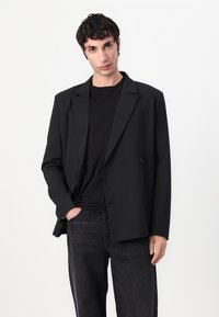 Schwarzes doppelt geknöpftes Blazer mit breiten Revers, strukturierten Schultern und einem lockeren Schnitt. Kombiniert mit einem schwarzen T-Shirt und dunklen Jeans.