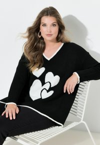 Černý oversized svetr s bílými srdíčkovými vzory a bílým lemováním podél výstřihu do V a manžet. Látka vypadá měkká a s jemnou texturou.