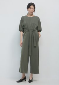 Bodysuit verde oliva con una vestibilità rilassata, maniche a palloncino corte, vita con fibbia, tasche laterali e pantaloni a gamba ampia. Abbinato a sandali neri con tacco largo.