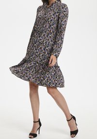 Abito floreale nero con maniche lunghe, orlo a volant, decorato con fiori viola e arancioni. Indossato con scarpe col tacco nere.