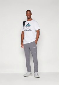 White t-shirt with blue Columbia logo, paired with light gray pants. Model wears gray sneakers, showcasing a casual outdoor look.