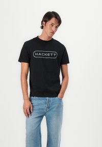 Juodas medvilnės marškinėlis su ryškiu pilku "HACKETT" logotipu. Su šviesiai mėlynomis džinsomis, demonstruojančiomis kasdienį įvaizdį.