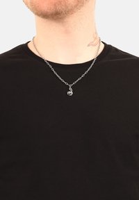 Collana in argento con un piccolo pendente decorativo, indossata sopra una maglietta nera, caratterizzata da un design a catena e una texture liscia.