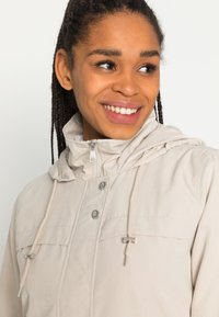 Chaqueta con capucha de color beige claro, con cremallera frontal, dos botones a presión y cordones ajustables en la capucha y la cintura. Tejido texturizado.