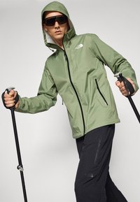 Light green waterproof jacket features a zippered front, adjustable hood, and two zippered side pockets. Model holds trekking poles.