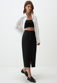 Jupe midi noire avec fente à l'avant, associée à un crop top noir et une chemise blanche à boutons. Des mocassins noirs complètent l'ensemble.