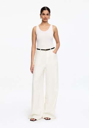 Žena v bielom tielku bez rukávov, biele široké nohavice s čiernym opaskom, zlaté kruhové náušnice, stojí s jednou rukou v vrecku.