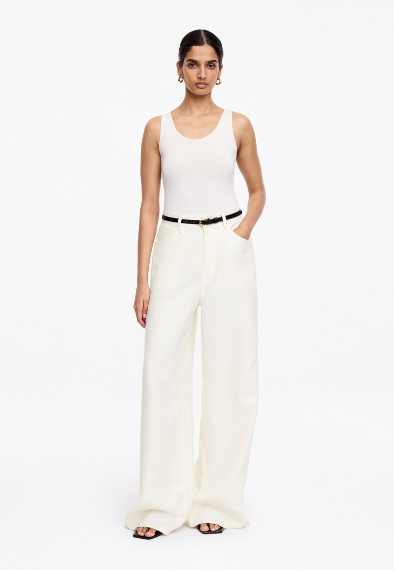 Femme portant un haut blanc sans manches, un pantalon blanc à jambes larges avec une ceinture noire, des boucles d'oreilles créoles dorées, debout avec une main dans la poche.