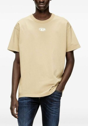 Uomo che indossa una t-shirt beige a maniche corte con piccolo logo bianco sul petto e jeans blu scuro, una mano in tasca.
