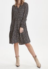 Blommig klänning med V-hals, långa ärmar och en vågig kjol. Svart bakgrund med färgglada blommor. Buren med beige ankelstövlar.