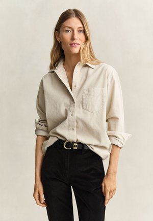 Femme aux longs cheveux blonds portant une chemise beige boutonnée avec les manches retroussées et un pantalon noir avec une ceinture noire, debout devant un fond clair.