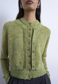 Γυναίκα που φοράει μια χνουδωτή πράσινη ζακέτα με κουμπιά και σκουλαρίκια με μαργαριτάρια, με τα χέρια στις τσέπες, μπροστά σε απ λό φωτεινό φόντο.