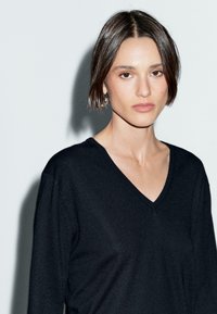 Una giovane donna con i capelli castani corti indossa un maglione nero a scollo a V, in piedi su uno sfondo grigio chiaro, con un'espressione seria.