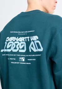 Felpa teal con collo rotondo, caratterizzata da una grande stampa grafica sul retro in testo bianco e azzurro chiaro, che cattura un'estetica di design retro.