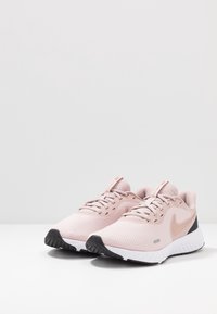 Paire de chaussures de course Nike rose pâle avec semelles intermédiaires blanches et semelles extérieures noires, posées sur une surface blanche avec un fond gris.