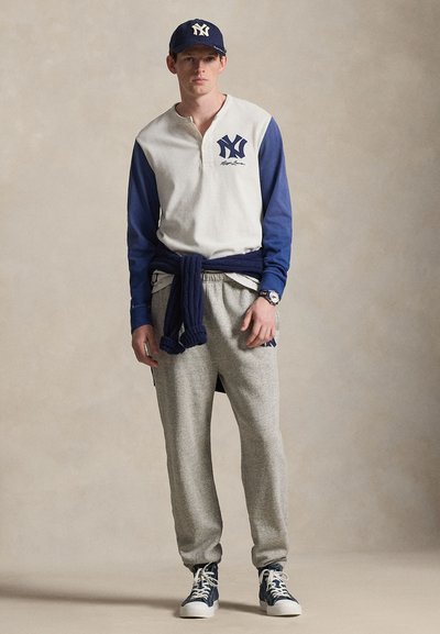 Fiatal férfi tengerészkék baseballsapkában, fehér és tengerészkék logós hosszú ujjú pólóban, szürke melegítőnadrágban, megkötött tengerészkék pulóverrel és tengerészkék magas szárú tornacipőben.