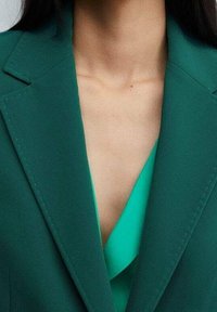 Blazer verde con rever a incavo, spalle strutturate e bordi netti, sovrapposto a una vivace maglia verde con scollo a V.