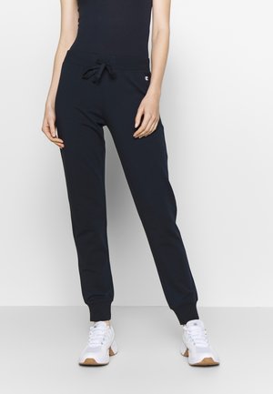 Woman wearing black drawstring sweatpants and white sneakers, standing against a plain white background.