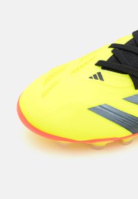 Botín de fútbol en color amarillo neón con cordones negros, cuerpo texturizado y rayas grises; presenta una suela de color rosa brillante y tacos moldeados para una mejor tracción.