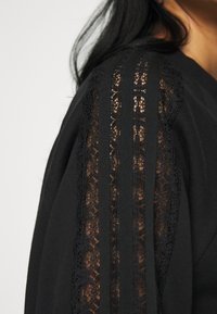 Svart topp med spetsdetaljer på ärmarna, med texturerad design och vertikala ränder som framhäver en blandning av material.