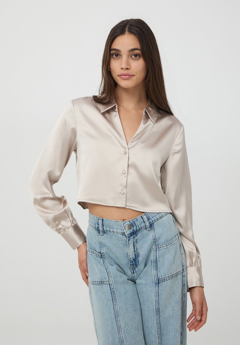 Blusa satinata cropped in beige chiaro con colletto, scollatura a v e chiusura frontale con bottoni. Abbinata a jeans azzurri sbiaditi a vita alta con tasche a toppa.