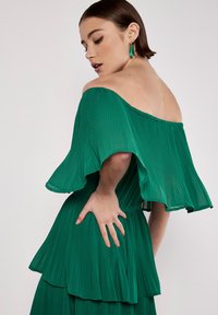 Grünes, plissiertes Off-Shoulder-Kleid mit weit ausgestellten Ärmeln und geschichtetem Rüschensaum. Glattes, leichtes Material mit einem dezenten Glanz.