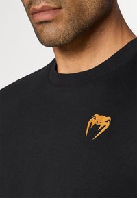 Svart bomull t-skjorte med rund hals og en liten oransje logo på venstre brystområde. Glatt tekstur, uformell design.