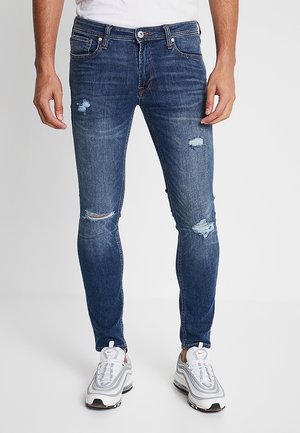 Mężczyzna w obcisłych niebieskich postrzępionych dżinsach i białych butach Nike, stojący na jednolitym jasnym tle.