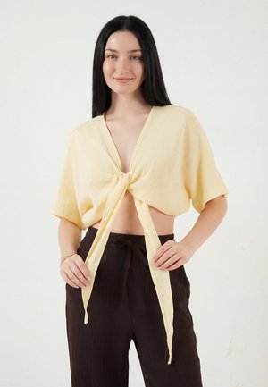 Giovane donna con capelli lunghi e neri, indossa una maglietta corta a maniche corte di colore giallo pallido annodata e pantaloni larghi marrone scuro, su sfondo semplice.