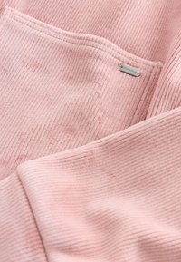 Gros plan sur un tissu côtelé rose avec une poche ornée d'une petite étiquette métallique portant la mention "DOWNTIME."
